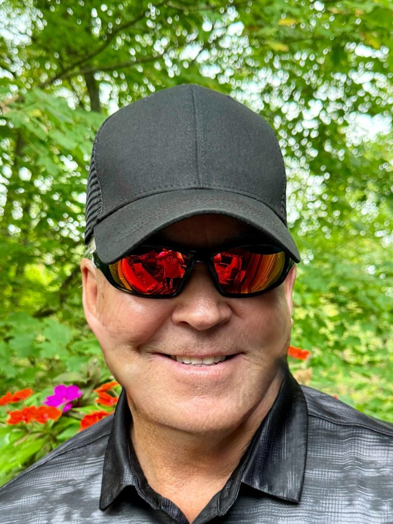 Smiling person wearing a black cap and sunglasses in a green outdoor setting.